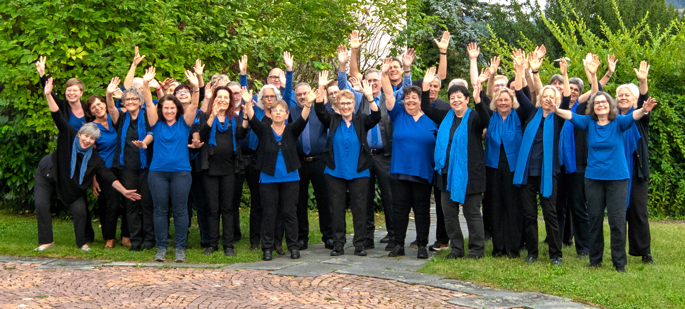 Die Mitglieder des Gospelchor Trimbach vor einem Hintergrund aus grünen Büschen. Sie zeigen ihre Freude am singen und werfen dabei ihre Hände hoch.